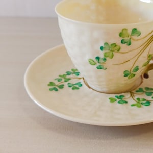 May include: A white teacup and saucer with a green shamrock design. The cup has a brown handle.