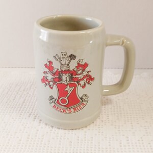 Peut inclure: Une chope à bière en céramique blanche avec un blason rouge et or représentant une clé et les mots "Beck's Bier".