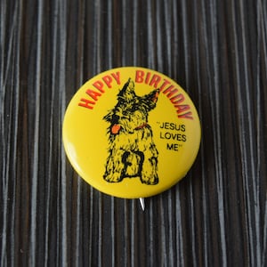 May include: Yellow button featuring a black and white illustration of a dog. The button reads "Happy Birthday" and "Jesus Loves Me".
