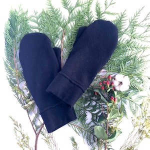 May include: A pair of black fleece mittens with a folded cuff. The mittens are lying on a bed of green pine branches and white cotton.