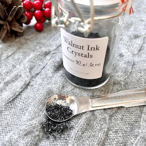 May include: A small glass jar filled with black walnut ink crystals. The jar has a white label that reads "Walnut Ink Crystals" and "Contents: 30 g / 6 tsp". The label also includes the name "Michiko Craft". A small metal measuring spoon with a black powder in it is next to the jar.
