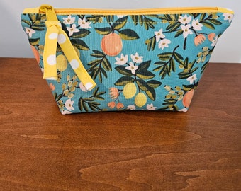 May include: A teal cosmetic bag with a yellow zipper and a matching polka-dot ribbon pull. The bag is decorated with a floral pattern featuring oranges, lemons, and white blossoms. The bag is sitting on a wooden surface.