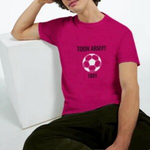 May include: A magenta t-shirt featuring the text "TOON ARMY!!" above a white and pink football graphic. Below the graphic is the number "1881". The shirt is worn by a person wearing dark green trousers.