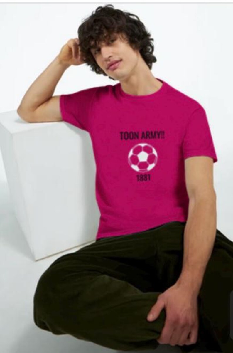 May include: A magenta t-shirt featuring the text "TOON ARMY!!" above a white and pink football graphic. Below the graphic is the number "1881". The shirt is worn by a person wearing dark green trousers.