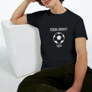 May include: Black t-shirt with the text "TOON ARMY!!" above a soccer ball graphic and the number "1881". The shirt is worn by a person sitting on a white cube, wearing dark green trousers.