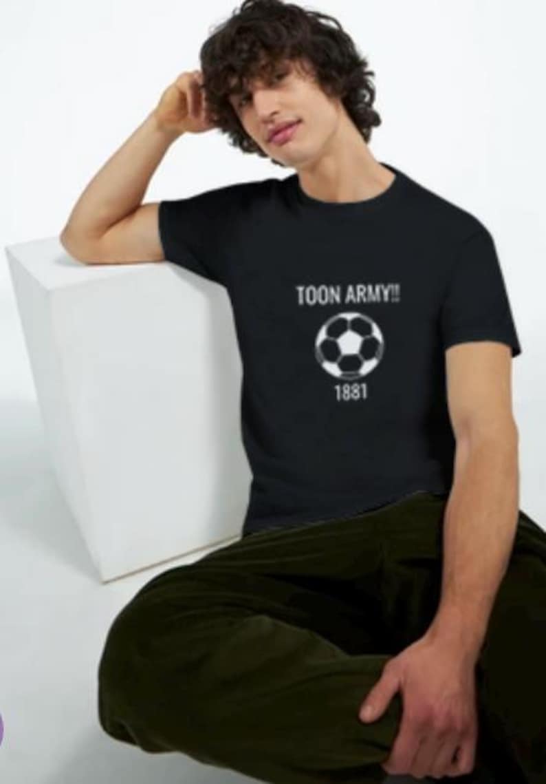 May include: Black t-shirt with the text "TOON ARMY!!" above a soccer ball graphic and the number "1881". The shirt is worn by a person sitting on a white cube, wearing dark green trousers.