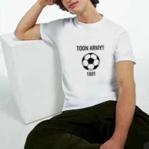 May include: White t-shirt featuring the text "TOON ARMY!!", a black and white football graphic, and the number "1881". The person is wearing dark green trousers.