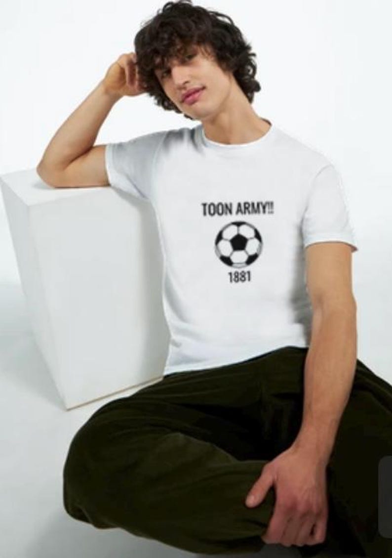 May include: White t-shirt featuring the text "TOON ARMY!!", a black and white football graphic, and the number "1881". The person is wearing dark green trousers.