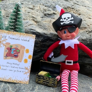 May include: A toy elf dressed as a pirate, wearing a black hat with a skull and crossbones, an eye patch, and holding a sword. Beside the elf is a treasure chest and a card that reads "Elf Treasure Island". The elf is wearing a red outfit with white and red striped legs.