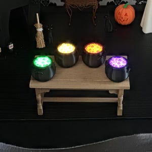 Four miniature black cauldrons sitting on a small wooden table, each filled with glowing bubbles in green, yellow, orange, and purple, with Halloween-themed decorations in the background.