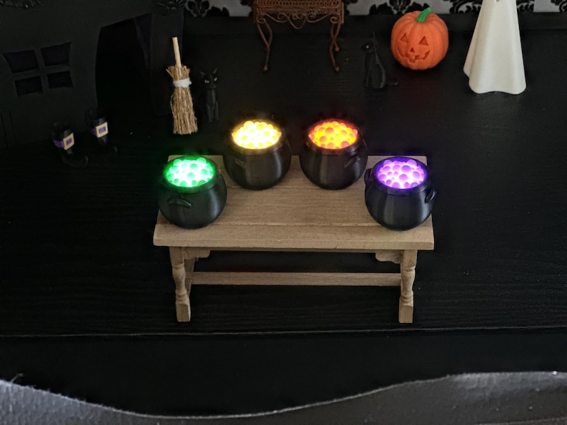 Four miniature black cauldrons sitting on a small wooden table, each filled with glowing bubbles in green, yellow, orange, and purple, with Halloween-themed decorations in the background.