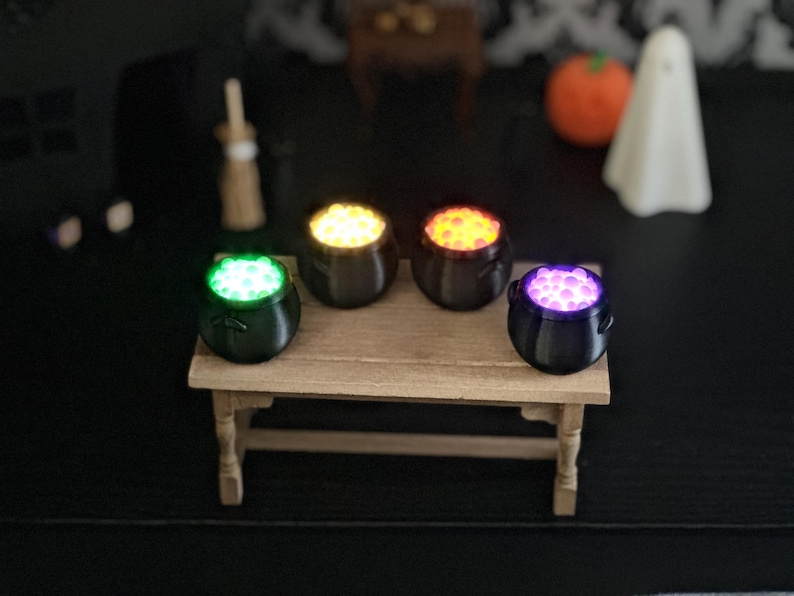 An elevated view of the four glowing miniature cauldrons arranged on a wooden miniature table in front of a Halloween diorama backdrop.