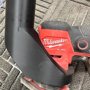 May include: A black 3D printed dust collection attachment for a Milwaukee FUEL brand sander. The sander is red and has a black handle. The sander is on a gray surface.