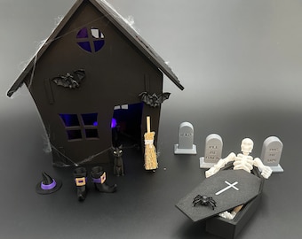 Peut inclure: Une maison d'Halloween noire avec une fenêtre éclairée, un chat noir, un balai et un chapeau de sorcière. Il y a aussi quatre petites pierres tombales avec le texte "RIP", "I'll Be Back" et "Dig Me Up". Un squelette blanc est allongé dans un cercueil noir avec une croix sur le dessus.