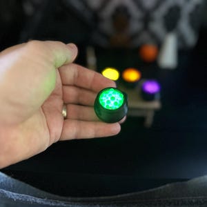 A wider shot of a hand holding up the glowing green miniature cauldron, with the table and other glowing cauldrons visible in the background.