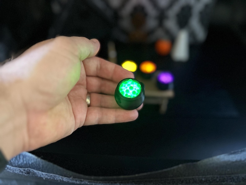 A wider shot of a hand holding up the glowing green miniature cauldron, with the table and other glowing cauldrons visible in the background.