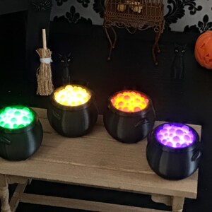 A close-up shot of the four miniature black cauldrons on a wooden table, featuring bright glowing bubble textures in green, yellow, orange, and purple.