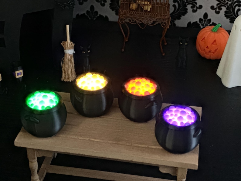 A close-up shot of the four miniature black cauldrons on a wooden table, featuring bright glowing bubble textures in green, yellow, orange, and purple.