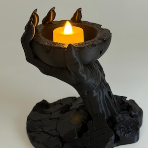 May include: A matte black candle holder shaped like a hand emerging from a cracked, textured base. The hand holds a circular bowl containing a small, glowing yellow LED tea light candle. The design features sharp, claw-like fingers.