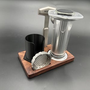 May include: AeroPress coffee maker set. The set includes a stainless steel coffee press, a black cup holder, a metal filter, and a stainless steel tamper. The items are displayed on a dark wood base. The AeroPress logo is visible on the tamper.