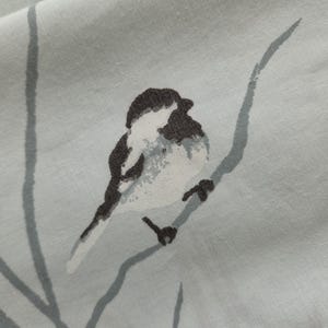 May include: A light gray fabric featuring a printed illustration of a small bird with black and white plumage perched on a thin, dark gray branch. The bird is depicted in a watercolor style with soft edges and visible brushstroke textures.