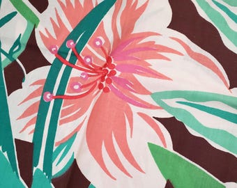 May include: Close-up of a fabric with a floral pattern. The design features a large, stylized flower with white petals edged in coral and pink. Green leaves and stems are interspersed against a dark brown background, creating a tropical theme.