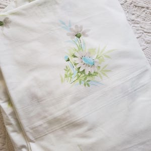 May include: White bed sheet with a floral pattern. The design features clusters of daisies in shades of gray, blue, and green. The sheet is folded, showing the pattern and the texture of the fabric. The background is a white, textured surface.