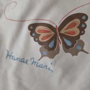 May include: A cream-colored fabric featuring a printed butterfly with dark brown wings, light blue accents, and orange details. A thin, curved orange line extends from the butterfly. The text "Hanae Mori" is printed in light blue script below.