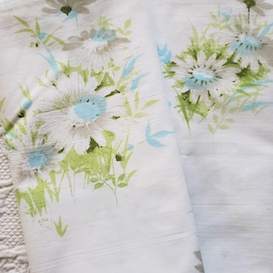 May include: White bed sheets with a floral pattern featuring large daisies in shades of white, gray, and light blue, with green leaves and accents. The sheets are folded and appear to be made of a soft fabric.