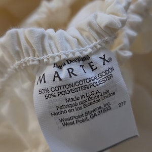 May include: A cream-colored fitted sheet with an elasticized edge and a white fabric tag. The tag reads: Extra Deeping MARTEX 50% COTTON/COTON/ALGODÓN 50% POLYESTER/POLIESTER Made in U.S.A. Fabriqué aux États-Unis Hecho en los Estados Unidos WestPoint Stevens, Inc.