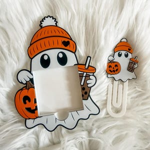 May include: Two Halloween-themed ghost-shaped accessories. One is a frame, the other a paperclip. Both ghosts wear orange beanies and hold pumpkins and bubble tea. The frame has a blank white space.