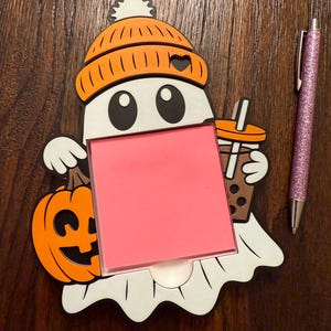 May include: A Halloween-themed memo holder shaped like a ghost wearing an orange beanie, holding a pumpkin and a bubble tea. A pink sticky note is in the center. A pink glitter pen is to the right.