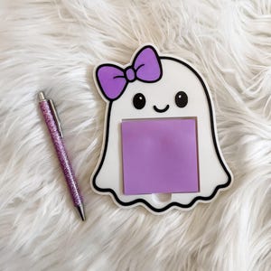 May include: A white ghost-shaped memo holder with a purple bow and a purple sticky note. A purple glitter pen is next to the memo holder. The memo holder has a black outline and a smiling face.