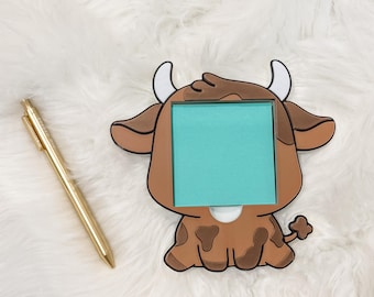 May include: A brown and white cow-shaped memo holder with a teal sticky note pad. The holder has a cartoon design with a white belly spot and small horns. A gold pen rests on the fluffy white surface.