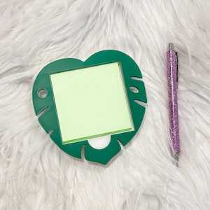 May include: A green, leaf-shaped memo holder with a light green sticky note pad. A purple glitter pen is next to the memo holder. The items are on a white, fluffy surface.