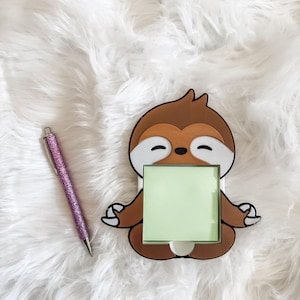 May include: A brown and white sloth-shaped memo holder with a light green square notepad. A pink glitter pen rests to the left of the memo holder. The sloth is in a seated yoga pose, with its arms and legs crossed.