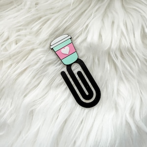 May include: A black paperclip with a coffee cup design. The cup is pink and mint green with a white lid and a white heart. The paperclip is a fun accessory for organizing papers or as a bookmark.