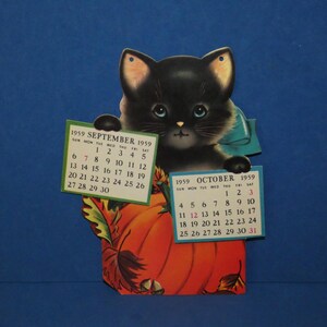 May include: A vintage die-cut paper calendar featuring a black kitten with blue eyes peeking over a large orange pumpkin. The kitten holds two small calendar pages for September 1959 and October 1959. The design includes autumn leaves and acorns.