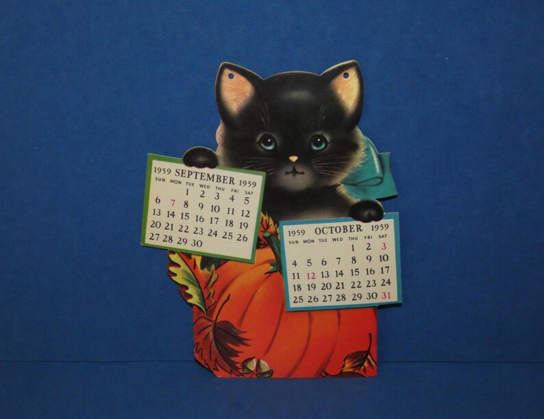 May include: A vintage die-cut paper calendar featuring a black kitten with blue eyes peeking over a large orange pumpkin. The kitten holds two small calendar pages for September 1959 and October 1959. The design includes autumn leaves and acorns.