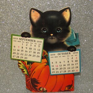 May include: A vintage 1959 calendar featuring a black kitten with blue eyes and a blue bow, holding two paper calendar pages for September and October. The kitten is positioned behind a large orange pumpkin decorated with autumn leaves and acorns.