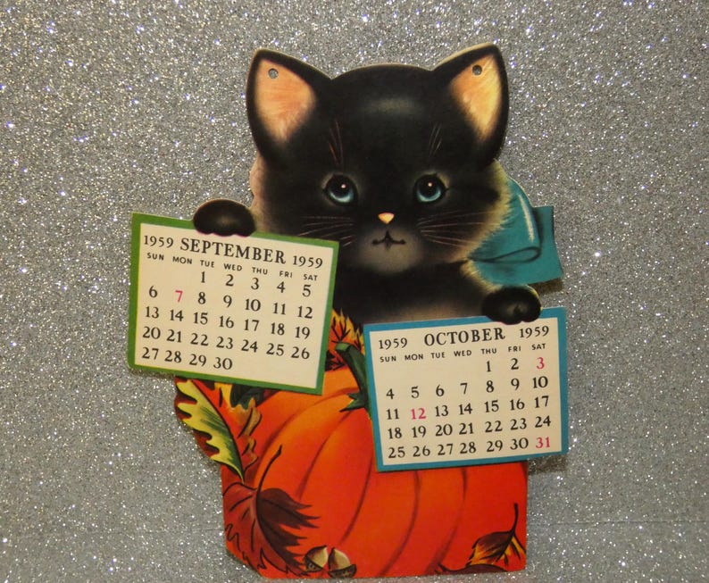 May include: A vintage 1959 calendar featuring a black kitten with blue eyes and a blue bow, holding two paper calendar pages for September and October. The kitten is positioned behind a large orange pumpkin decorated with autumn leaves and acorns.