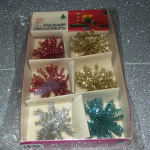 May include: A rectangular box of Package Decorations containing six star-shaped tinsel ornaments in red, gold, silver, and blue. The packaging features the text Package Decorations and the number 1636. The items are encased in clear plastic wrap.