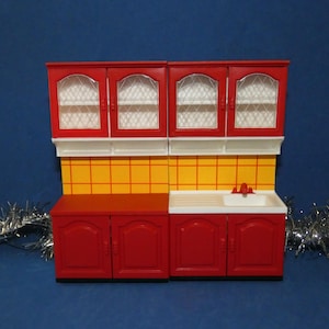 May include: A red and white toy kitchen set with upper cabinets, a yellow tiled backsplash, and lower cabinets with a sink. The upper cabinets have arched windows. The set is on a blue background.