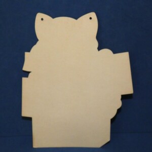 May include: A light-colored, flat wooden cutout shaped like a cat head with pointed ears. The object features two small circular holes at the top for hanging and has a smooth, unfinished surface with a distinct silhouette and straight edges.