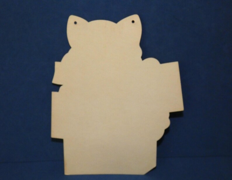 May include: A light-colored, flat wooden cutout shaped like a cat head with pointed ears. The object features two small circular holes at the top for hanging and has a smooth, unfinished surface with a distinct silhouette and straight edges.