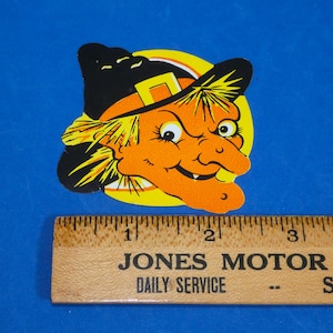 May include: A die-cut paper witch decoration featuring an orange face, yellow hair, and a black hat with a yellow buckle, positioned above a wooden ruler showing a width of approximately 3.5 inches for the witch graphic.