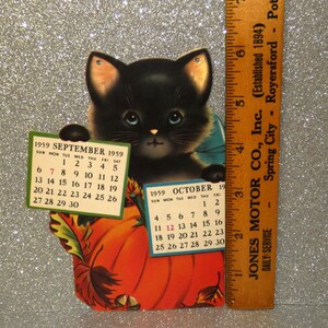 May include: A vintage 1959 calendar featuring a black kitten sitting behind a large orange pumpkin. The kitten holds two small calendar pages for September and October 1959. A wooden ruler placed next to the item shows a height of 6 inches.