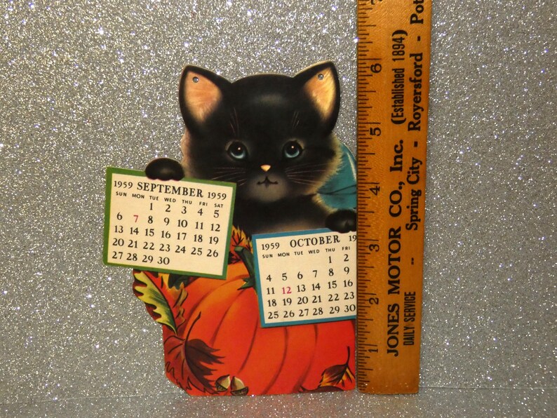 May include: A vintage 1959 calendar featuring a black kitten sitting behind a large orange pumpkin. The kitten holds two small calendar pages for September and October 1959. A wooden ruler placed next to the item shows a height of 6 inches.