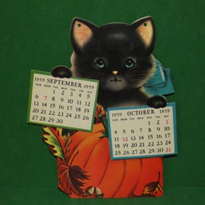 May include: A vintage 1959 wall calendar featuring a black kitten with blue eyes peeking over a large orange pumpkin. The kitten holds two paper calendar pages for September and October 1959. The design includes autumn leaves and acorns.