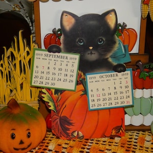 May include: A vintage 1959 calendar featuring a black kitten illustration holding two small cards for September and October. The kitten sits atop a large orange pumpkin surrounded by autumn leaves and acorns, with a jack-o'-lantern nearby.